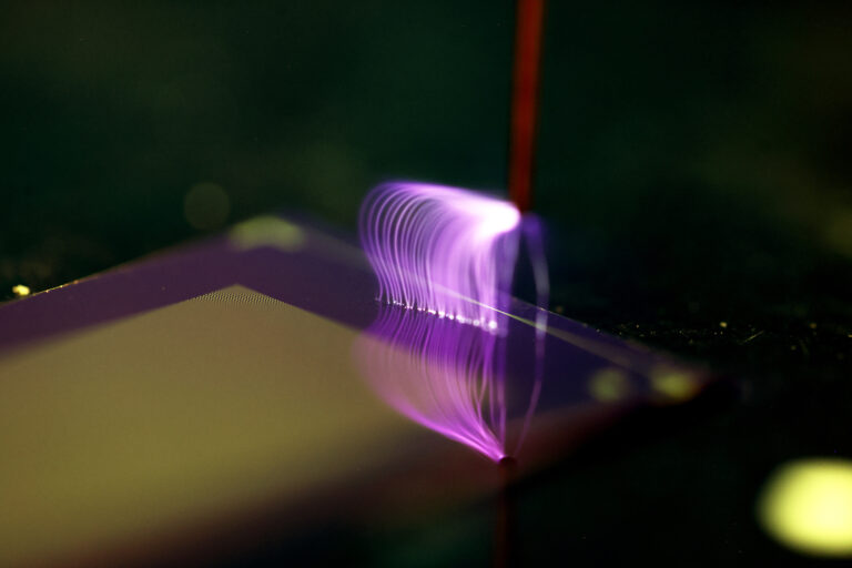 Bright purple, lightning-like plasma arcs in thin filaments from the tip of the metal wand onto the blue part of the chip, resembling a waterfall. The plasma stands in stark contrast with the dark background and illuminates the wand, which rises like a stem behind the arcs.