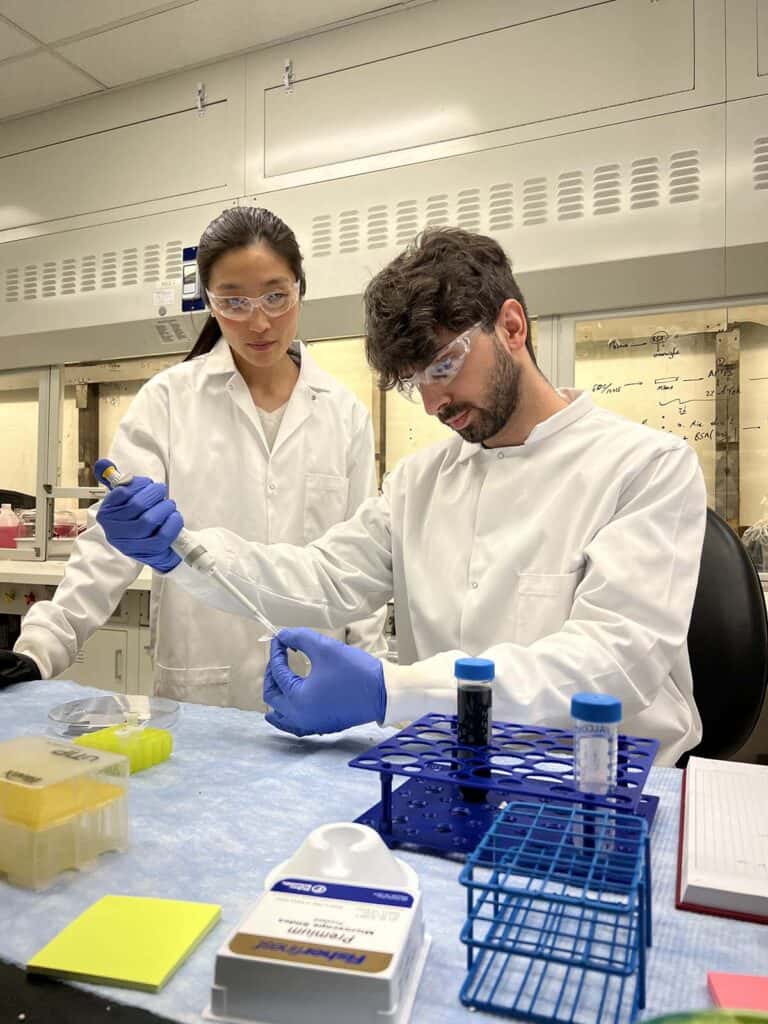 Two scientists wearing white coats and safety glasses work in front of a lab bench. One scientist is using a mechanical pipette to add liquid into a small test tube. Other test tubes arranged on the bench hold a black liquid.